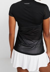 Svart kortärmad topp med genomskinlig ryggdesign, med rund halsringning och Lacoste-logotyp på baksidan. Matchad med en vit plisserad kjol.