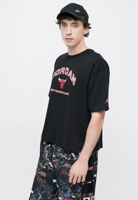 Μαύρο βαμβακερό T-shirt με άνετη εφαρμογή, με κόκκινο κείμενο "JORDAN" και λογότυπο ταύρου στο στήθος. Συνδυάζεται με σορτς με σχέδια.