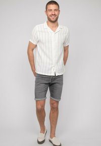 Chemise blanche à manches courtes avec des rayures noires, associée à un short en denim gris retroussé et des chaussures échancrées beiges. Tissu léger et texturé.
