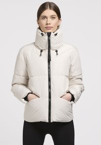 Doudoune de couleur claire avec un col haut, une fermeture éclair noire et des cordons réglables. Dotée d'un design matelassé et de poches latérales.