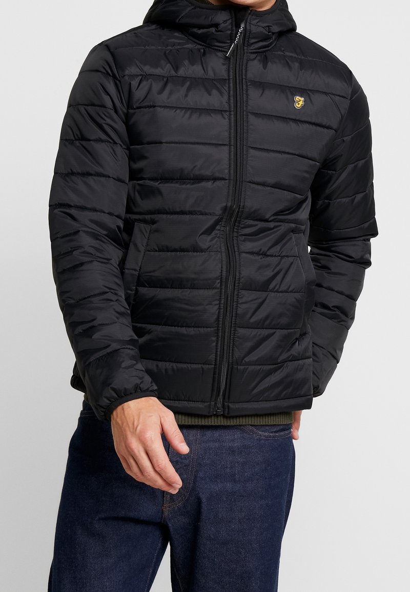 Veste matelassée noire avec capuche, présentant des coutures horizontales, une fermeture éclair frontale et un petit logo jaune sur la poitrine. Finition texturée.