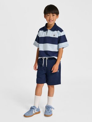 Young boy stands smiling, wearing a navy and light blue striped polo shirt, navy shorts, white socks, and light blue sneakers.