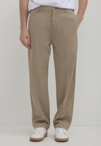 Pantaloni beige con una vestibilità comoda, caratterizzati da una parte anteriore piatta, tasche laterali e chiusura con bottone. Indossati con sneaker bianche.