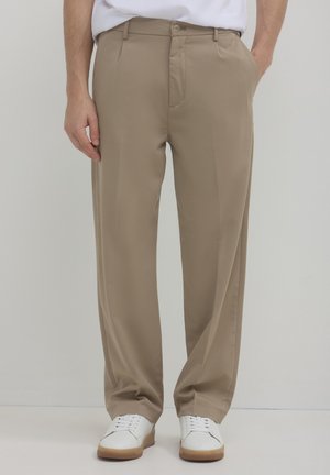 Beige Hosen mit lockerem Schnitt, mit flacher Vorderseite, Seitentaschen und Knopfverschluss. Getragen mit weißen Sneakers.