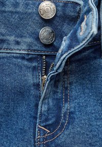 Los jeans de denim azul presentan botones metálicos plateados y una cremallera metálica. La tela tiene un acabado texturizado con costuras visibles y acentos desgastados.