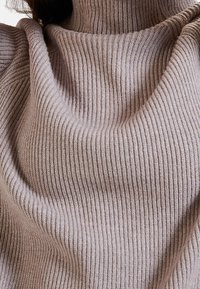 Hedvábný rolák v měkké béžové látce, který má strukturovaný vzor a uvolněný střih. Mezi detaily nalezneme vysoký límec a volně splývající výstřih.