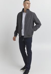 Jachetă gri cu fermoar și guler înalt, asortată cu un tricou alb și pantaloni bleumarin. Tenisi negri completează ținuta.