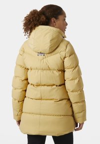 Žlutá puffer bunda s kapucí, prošívaný design a šedý akcent loga na zadní straně. Bunda má hladkou texturu a uvolněný střih.