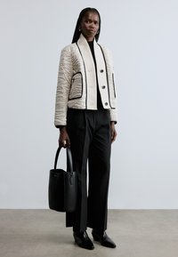 Sieviete ar pīnētiem matiem, tērpusies krēmkrāsas dūraiņu jakā ar melnām apmalēm, melnās biksēs, melnos zābakos un turot melnu ādas ikdienas somu.