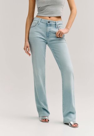 Femme portant un jean taille haute bleu clair avec des détails en strass et des talons ouverts argentés, debout avec une main sur la hanche.