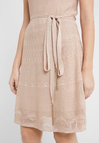 Robe en maille rose clair avec un motif texturé et une ceinture nouée à la taille. Dotée d'accents dorés aux extrémités de la ceinture.