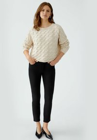 Beige gebreide trui met een textuurpatroon van golven, zwarte skinny jeans en zwarte puntenballetsleutels, gedragen door een persoon die staat met de handen in de zakken.