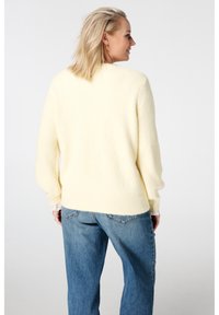 Geel gebreide trui met een zachte textuur en geribde zoom, gecombineerd met lichtblauwe spijkerbroek. De trui heeft een losse pasvorm en lange mouwen.