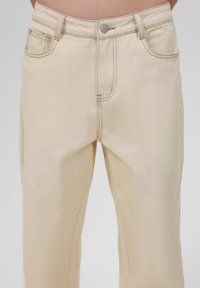 Beige denim pants with a straight-leg design, featuring a front button closure, five pockets, and contrasting stitching details.