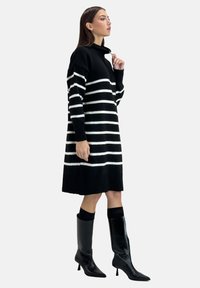 Robe tricotée à rayures noires et blanches avec un col montant, des manches longues et des poignets côtelés, associée à de hautes bottes en cuir noir.