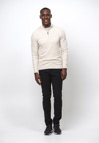 Maglione beige chiaro con collo con zip, tessuto strutturato; abbinato a pantaloni neri e sneaker scure; modello in piedi contro uno sfondo semplice.