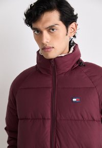 Bordeaux gefütterte Jacke mit hohem Kragen, Reißverschluss und Fleece-Futter. Verfügt über ein kleines Logo-Patch auf der Brust zur Markenidentifikation.