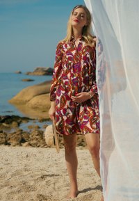 F�ärbiger, gemusterter Kleid mit Paisleymuster in Rot-, Grün- und Violetttönen. Leichter Stoff, lange Ärmel und eine Knopfleiste. Hält einen Strohhut.