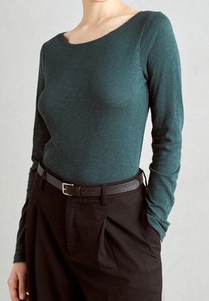 Kvinde iført en mørk teal langærmet top, stoppet ned i højtaljede sorte bukser med læg, accessorized med et sort bælte.