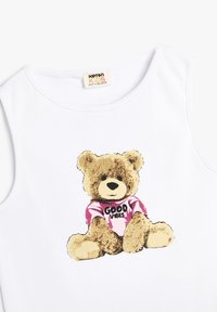 Camiseta sin mangas blanca de canalé con un gráfico de un oso que lleva una camisa rosa con el texto "BUENAS VIBRACIONES". Material de algodón con escote redondo.