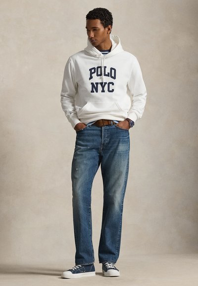 Άνδρας που φορά λευκό φούτερ με κουκούλα και την επιγραφή «POLO NYC», μπλε τζιν, μπλε αθλητικά παπούτσια, με τα χέρια στις τσέπες, κοιτάζοντας προς τα κάτω αριστερά μπροστά σε μπεζ φόντο.