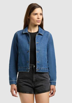 Vrouw met steil bruin haar, gekleed in een blauw spijkerjasje, zwart shirt en zwarte shorts met hoge taille, kijkt naar de zijkant.