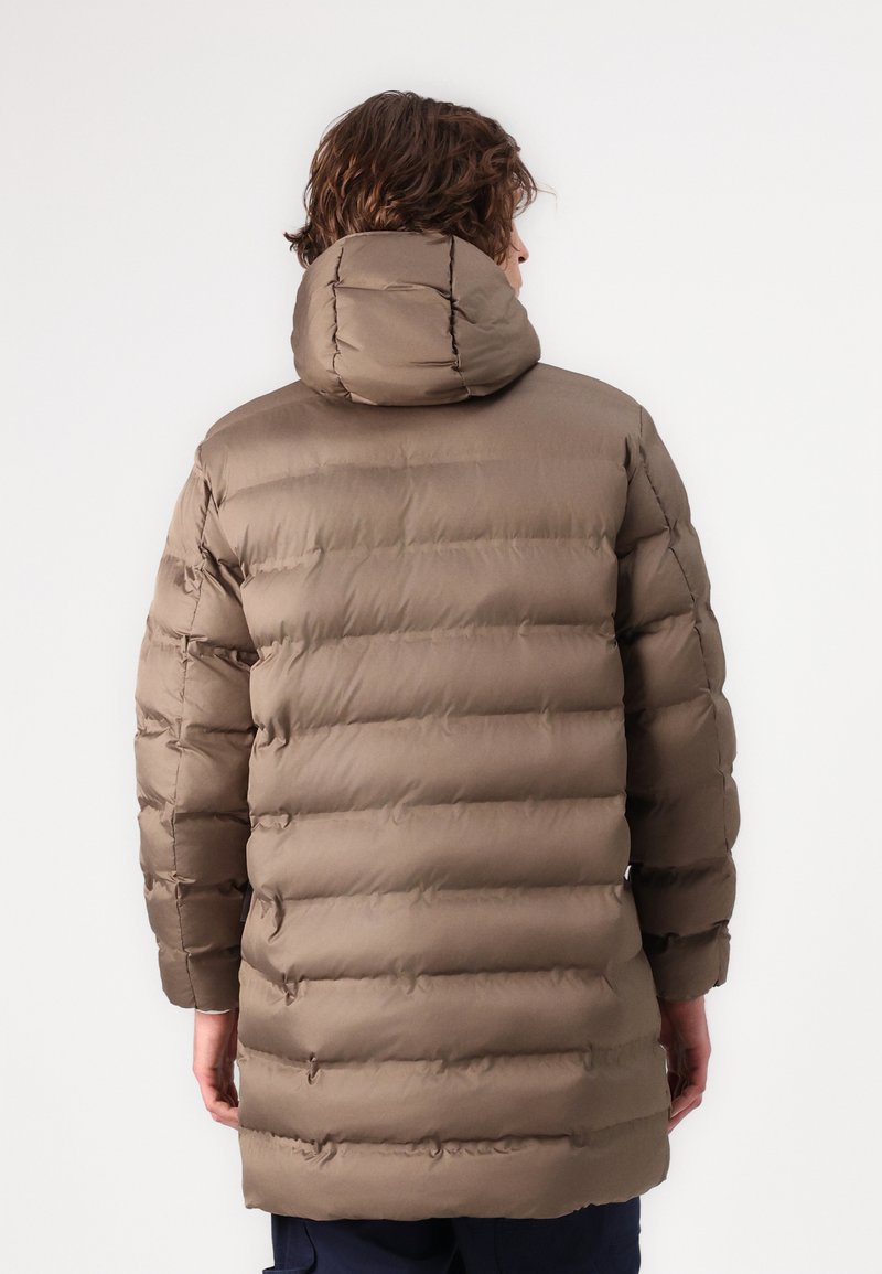 Person mit lockigem Haar, der eine lange, gesteppte braune Daunenjacke mit Kapuze trägt und von einem schlichten weißen Hintergrund abgewandt ist.