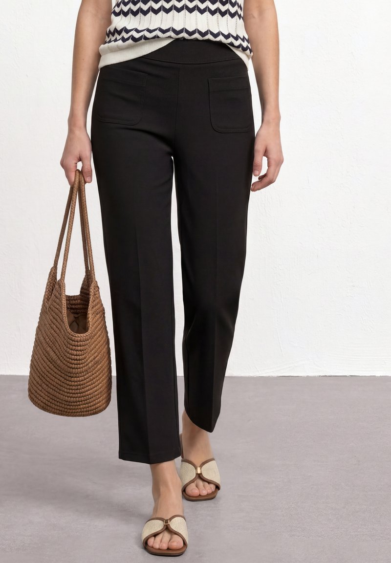 Woman wearing black pants, a patterned sleeveless top, beige sandals, holding a brown woven tote bag against a plain background.
