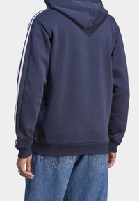 Tummansininen huppari, jossa teksturoitu pinta, valkoiset raidat hihoissa ja huppu. Suunniteltu rentoon istuvuuteen ja V-mallinen takaosa.