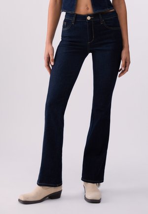 Žena v tmavomodrých džínsach bootcut strihu, skrátenej denimovej veste a bielych kozačkách na opätku stojaca na jednoduchom svetlom pozadí.