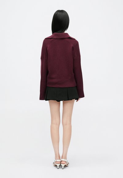 Bordó kötött pulóver gallérral, bordás szegéllyel és leengedett vállakkal, fekete plisszírozott szoknyával és fehér sarkú szandállal párosítva.