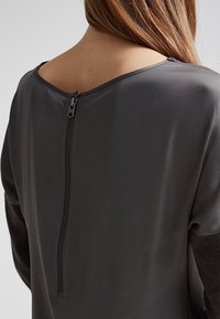 Blouse grise avec une texture satinée lisse, un col rond et une fermeture éclair visible dans le dos, avec des manches longues en tissu contrastant.