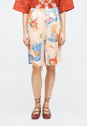 Σορτς μέχρι το γόνατο με πολύχρωμες εκτυπώσεις θαλάσσιας ζωής, φορεμένα με κόκκινα φλατ παπούτσια με κορδόνια και πορτοκαλί τοπ με λευκό σχέδιο κοραλλιού.