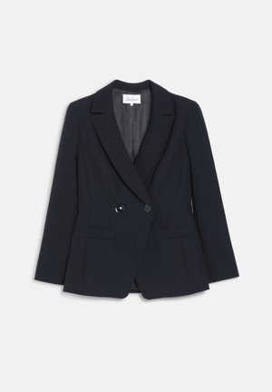 Kvinde iført en sort skræddersyet dragt med en mønstret sort-hvid bluse, der holder i reverserne på sin blazer og står mod en ensfarvet baggrund.