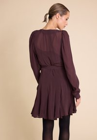 Kort lilla kjole laget av lett stoff med lange ermer, en figursydd midje og en utflaring nederdel. Plisserte detaljer fremhever bevegelse.