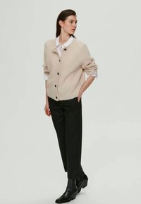 Kvinde iført en beige knaplukket cardigan, hvid skjorte med krave, sorte bukser og sorte ankelstøvler, stående med den ene hånd i lommen.