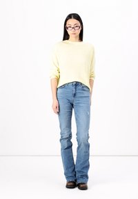 Svetlo rumena pletena jopica, modri raztrgani flared jeans in temno rjave čevlje. Model stoji pred enobarvno belo ozadje.