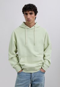 Young man with curly dark hair wears light green hoodie and blue jeans, standing with hands in pockets against plain background.