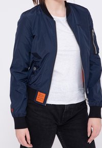 Veste bomber bleu marine avec fermeture éclair à l'avant, poignets et ourlet côtelés. Présente un patch orange sur le côté et une poche zippée sur la manche.