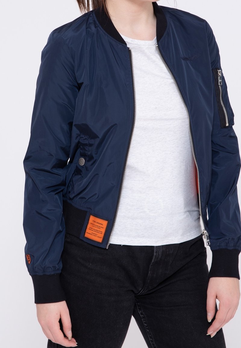 Veste bomber bleu marine avec fermeture éclair à l'avant, poignets et ourlet côtelés. Présente un patch orange sur le côté et une poche zippée sur la manche.