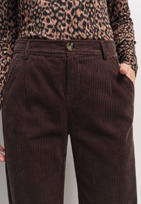 Brązowe spodnie korkowe z pionowym prążkowaniem, z zapięciem na guzik i kieszeniami bocznymi. Noszone z wzorzystą górą.