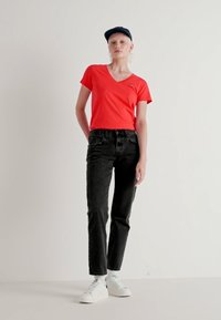 Rød V-hals t-skjorte sammen med høy belte svart jeans og hvite sneakers. Modellen har på seg en marineblå caps og står mot en nøytral bakgrunn.