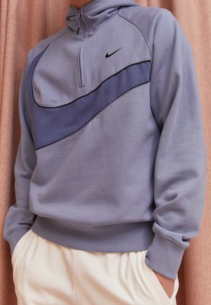 Henkilö, joka käyttää vaaleanpunaista Nike-pulloveria, jossa on tummanvioletti diagonaalinen raita ja puolivetoketju, yhdistettynä kermaisenvärisiin housuihin.