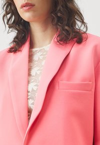 Giacca rosa con rever a intaglio, taschino sul petto e texture morbida, indossata sopra una blusa floreale trasparente.