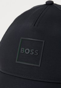 Black cap made of smooth fabric, featuring a square patch with "BOSS" in black text outlined by a green border. Simple design, no patterns.