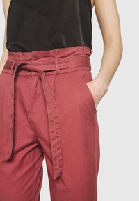 Bordeaux hoge taille broek met een brede riem, zijzakken en een gladde textuur, aangevuld met gestikte details langs de naden.