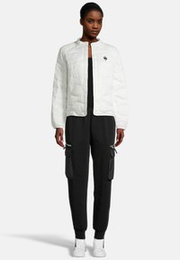 Veste matelassée blanche avec un col arrondi, associée à un pantalon cargo noir et des baskets blanches. Détails notables : poches zippées.