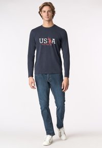 T-shirt a maniche lunghe navy con grafica di testo "USA" in bianco e rosso; abbinato a jeans di denim blu e sneakers bianche, in piedi su uno sfondo semplice.