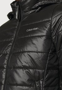 Sort pufferjakke lavet af glat materiale, med quiltet design, lynlås foran og hætte med justerbare snore. Calvin Klein-logoet vises.