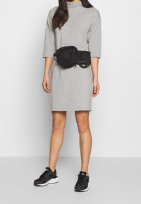 Černá ledvinka se dvěma přihrádkami, nošená přes šedé šaty. Oblečení je doplněno černými teniskami s bílými detaily.